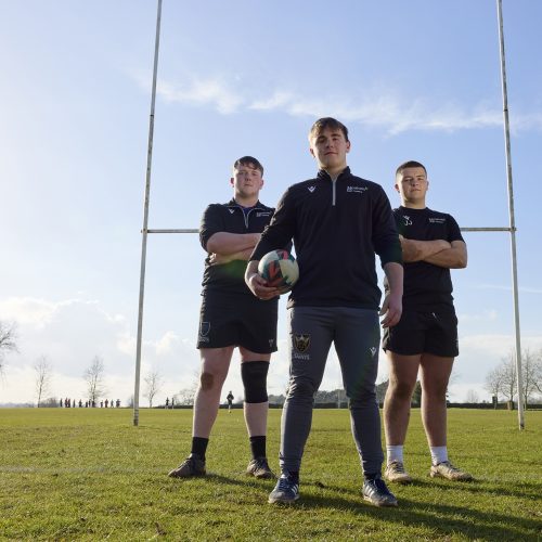 Three students wearing rugby kit standing in front of rugby posts